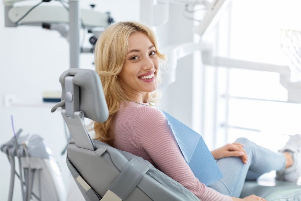Smiling and relaxed young woman sitting at dental chair in modern clinic, waiting for her dentist